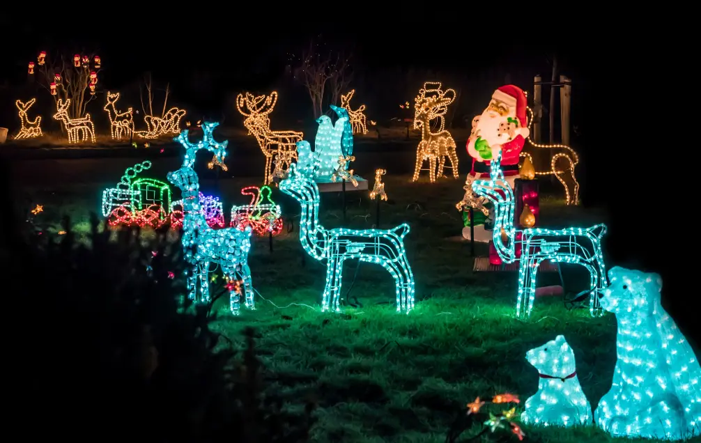 A full-yard Aldo Unlimited holiday lighting display — lit reindeer, Santa, and trees glowing at twilight.